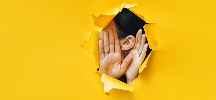 Eine Frau hält ihr Ohr durch ein Loch in gelber Papierwand