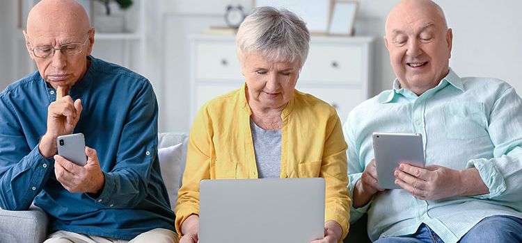 Zwei Rentner und eine Rentnerin auf Sofa, sie checken ihre Laptops und Smartphones