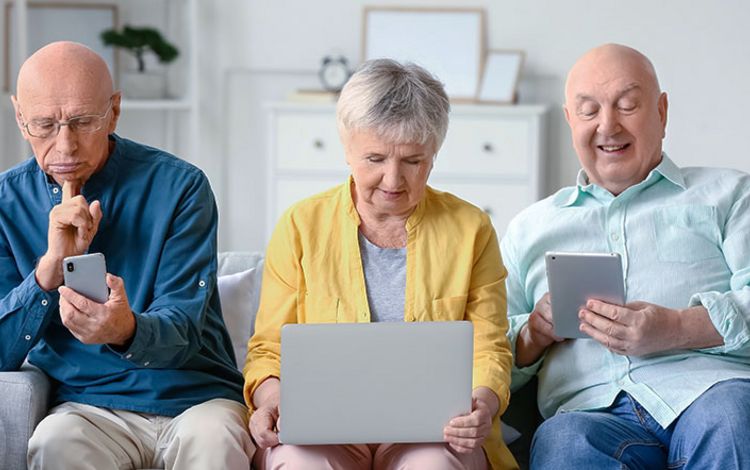 Zwei Rentner und eine Rentnerin auf Sofa, sie checken ihre Laptops und Smartphones