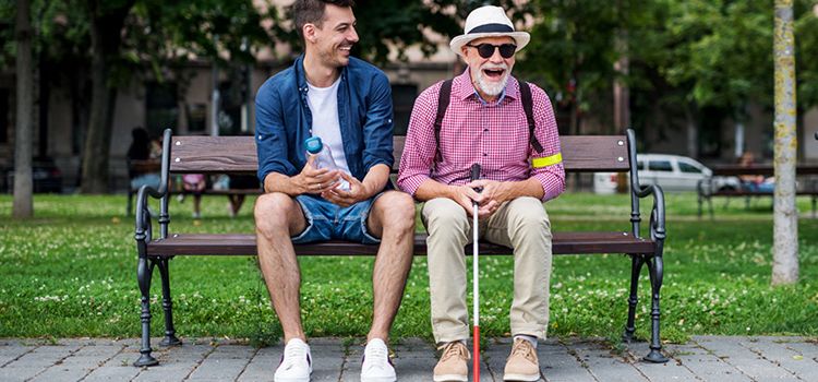 Alter blinder Mann auf einer Parkbank lacht zusammen mit einem jungen Mann