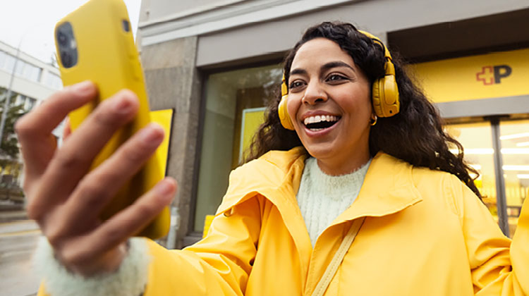 Junge Frau mit gelbem Smartphone und gelber Jacke lacht und macht ein Selfie