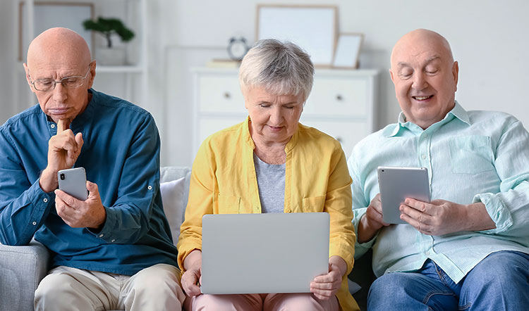 Zwei Rentner und eine Rentnerin auf Sofa, sie checken ihre Laptops und Smartphones